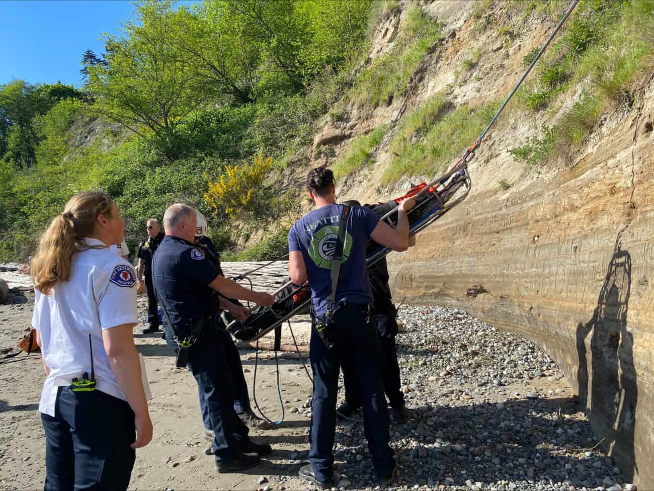 Firefighters lower a rescued hiker down a sandy embankment to the beach.